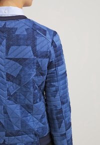 Gequiltete blaue Jacke mit einem geometrischen Muster, in verschiedenen Blautönen und mit einer glatten, strukturierten Oberfläche.