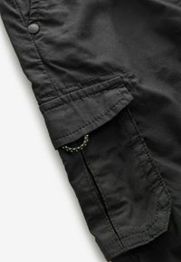 Close-up of black fabric pants pocket with flap secured by stitching and a small loop of black and green braided cord visible underneath.