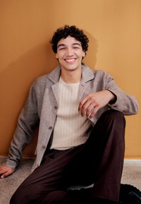 Bărbat tânăr cu păr creț, așezat pe podea, purtând cămașă bej, jachetă gri și pantaloni închiși la culoare, zâmbind pe fundalul unui perete galben muștar.