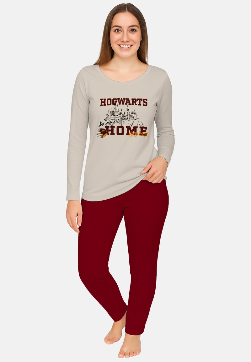 Giovane donna sorridente, che indossa una maglietta a maniche lunghe beige con la scritta "Hogwarts è casa mia" e un grafico del castello, abbinata a pantaloni rossi, a piedi nudi.