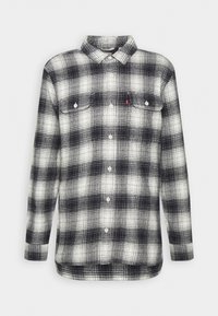 Chemise à carreaux noir et blanc à manches longues avec boutons, deux poches poitrine et un col, présentée sur un fond uni.