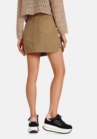 Gonna di denim color khaki con taglio dritto e dettaglio con spacco laterale, abbinata a scarpe sportive nere e rosa con suola bianca spessa.