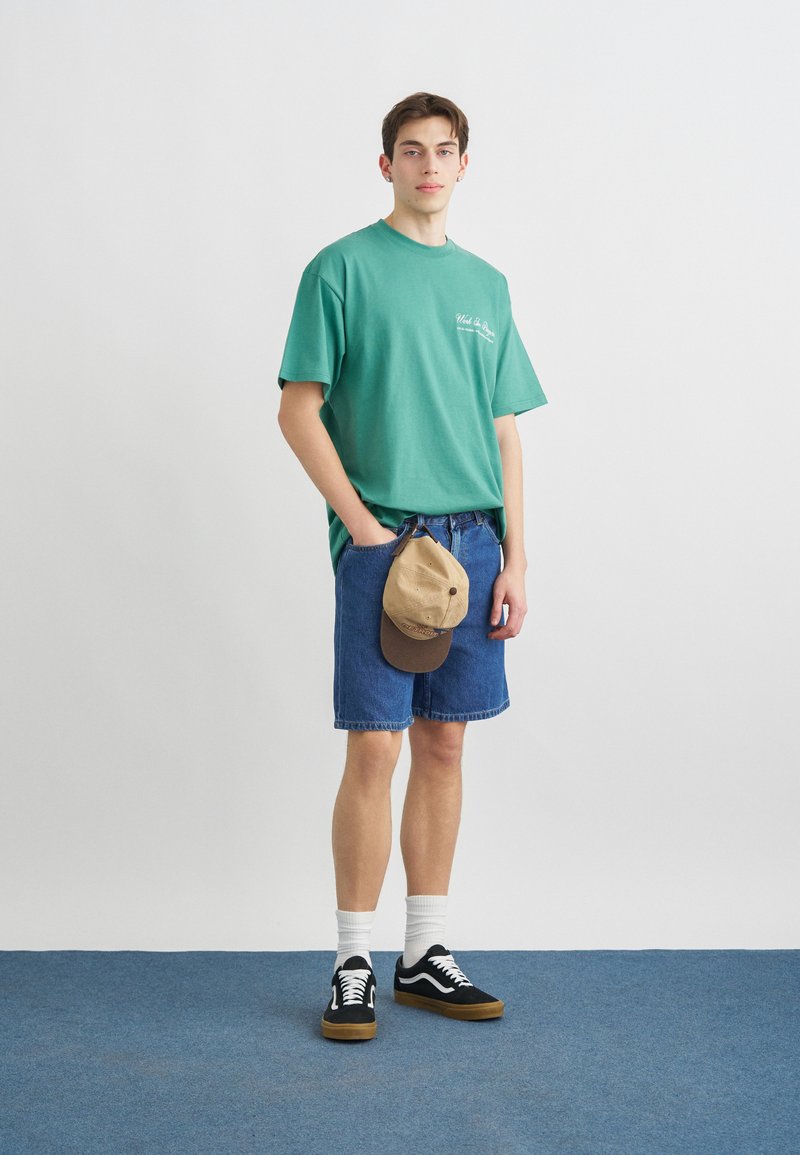Joven de pie con las manos en los bolsillos, vistiendo una camiseta verde, pantalones cortos de mezclilla azul, calcetines blancos, zapatillas negras y sosteniendo una gorra beige de béisbol.