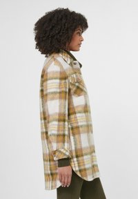 Lang flanellen shirt in een gele, groene en witte ruitpatroon. Zacht materiaal, met knopen aan de voorkant en twee borstzakken. Zijkant toont de lengte.