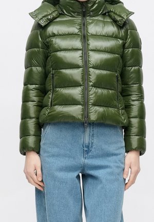 Femme portant une doudoune verte brillante avec fermeture éclair et deux poches avant, associée à un jean taille haute bleu clair.