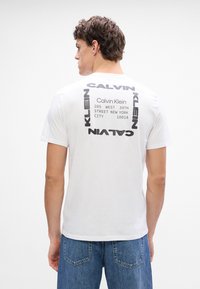 Tricou alb din bumbac, cu un design imprimat pe spate, având text negru îndrăzneț, inclusiv numele brandului și detaliile adresei.