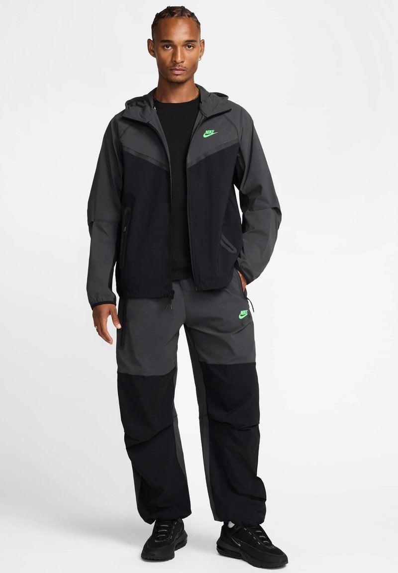 Chaqueta y pantalones Nike en negro y gris con capucha, bolsillos con cremallera y acentos de logo verde. Tejido ligero y transpirable, y corte holgado.