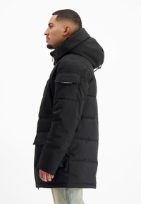 Man in profiel met een zwarte gewatteerde winterjas met een capuchon en klepzakken, gecombineerd met lichtblauwe jeans.