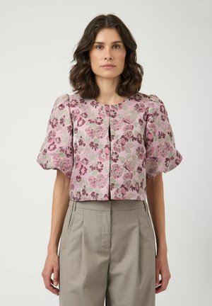 Õlgadeni laineliste juustega naine, kes kannab roosat lillelist pluusi, millel on laiad varrukad, ja kõrge vöökohaga beežid pükse, seisab ühevärvilise tausta ees.