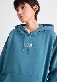 Sweatshirt à capuche couleur bleu sarcelle, en tissu doux, avec une poche kangourou et un logo brodé blanc sur la poitrine.