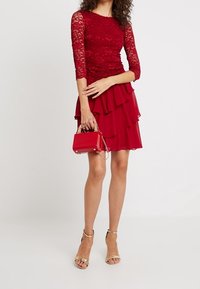 Top rosso in pizzo a maniche lunghe sopra una gonna rossa a strati, abbinato a una piccola borsa rossa e sandali con cinturino metallico. Texture liscia, silhouette aderente.