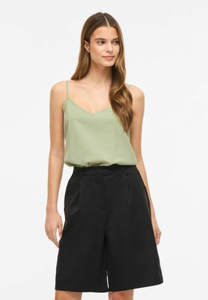 Naine pikkade pruunide juustega, kes kannab helesinise varrukateta topi ja musti põlveni ulatuvaid lühikesi pükse, seisab ühevärvilise tausta ees.