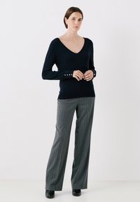 Marineblauer V-Ausschnitt-Pullover aus weichem Strick mit langen Ärmeln. Mit goldenen Knopfakzenten an den Bündchen. Kombiniert mit grauen, gestreiften, weit geschnittenen Hosen.