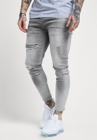 Jeans ajustados gris con detalles deshilachados en las rodillas y los muslos, con una textura suave. Combinados con zapatillas atléticas blancas, cortas por encima del tobillo.