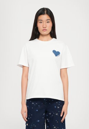 Camiseta blanca de manga corta con un parche de corazón de denim azul en el lado izquierdo del pecho con el texto "WE GO" debajo. Ajuste informal, material de algodón.