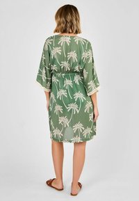 Grünes, transparentes Kleid mit einem Palmbaum-Print, einem Kordelzug in der Taille und Spitzensaum an den Ärmeln. Flache Sandalen runden das Outfit ab.