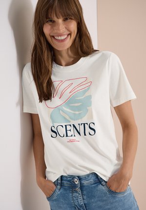 Smilende kvinde med brunt hår iført en hvid T-shirt med blåt og rødt løvdesign og teksten "SCENTS" samt blå jeans med hænderne i lommerne.