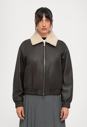 Woman wearing a dark brown leather jacket with a beige shearling collar and a gray pleated skirt, facing forward against a plain background.