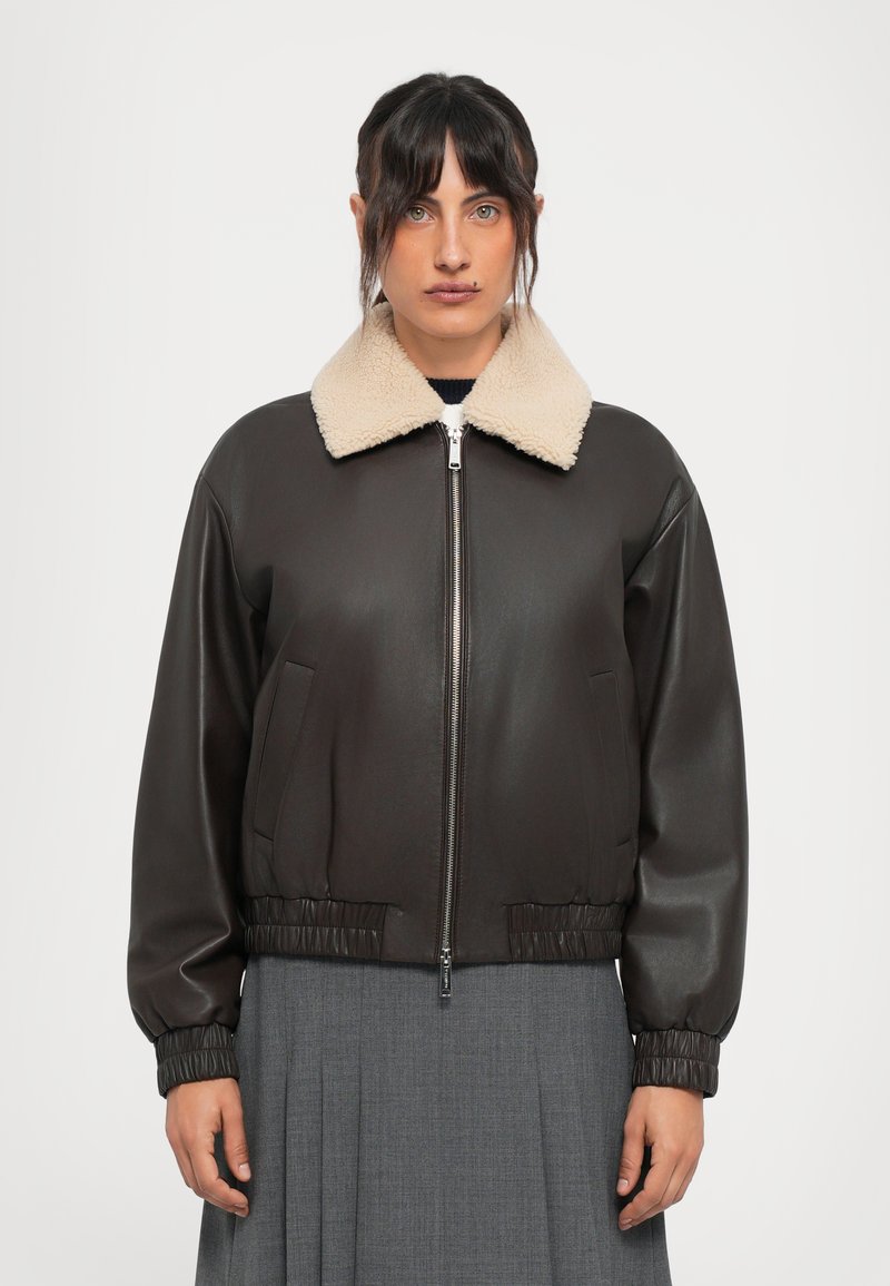 Woman wearing a dark brown leather jacket with a beige shearling collar and a gray pleated skirt, facing forward against a plain background.
