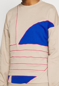 Krémová mikina s modrými a růžovými geometrickými vzory a detaily v horizontálních pruzích, vyrobená z měkké tkaniny s volným střihem.