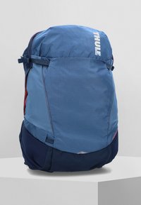 Mochila Thule azul con un tejido texturizado, cierre de cuerda en la parte superior, bolsillo lateral rojo y panel inferior en azul marino. Incluye correas ajustables.