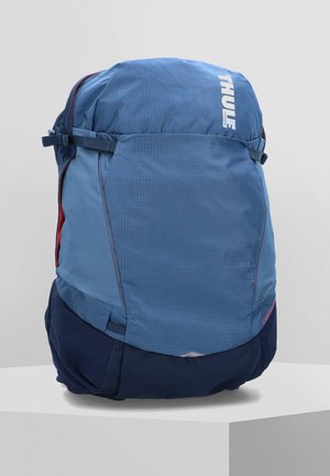 Mochila Thule azul con un tejido texturizado, cierre de cuerda en la parte superior, bolsillo lateral rojo y panel inferior en azul marino. Incluye correas ajustables.