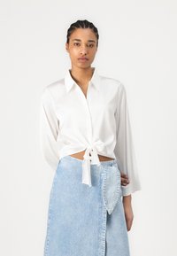 Blusa bianca a maniche lunghe con nodo legato in vita, abbinata a una gonna in denim azzurro chiaro con una tasca unica a forma di foglia.