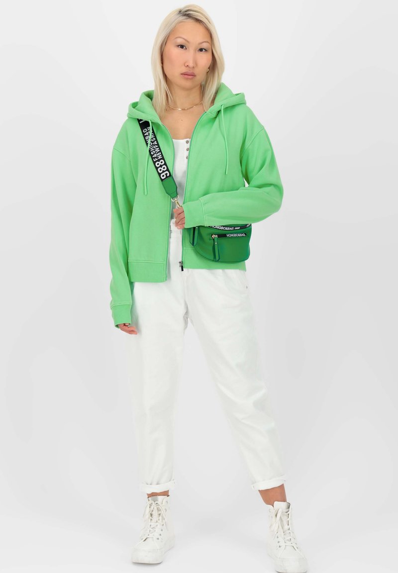 Femme portant un sweat à capuche vert, un pantalon blanc, des baskets montantes blanches et un sac bandoulière vert avec une sangle noire, se tenant contre un fond uni.