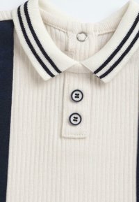 Polo à col avec texture côtelée, corps crème et rayures bleu marine sur les épaules. Disponible avec deux boutons foncés et une fermeture pression au niveau du col.