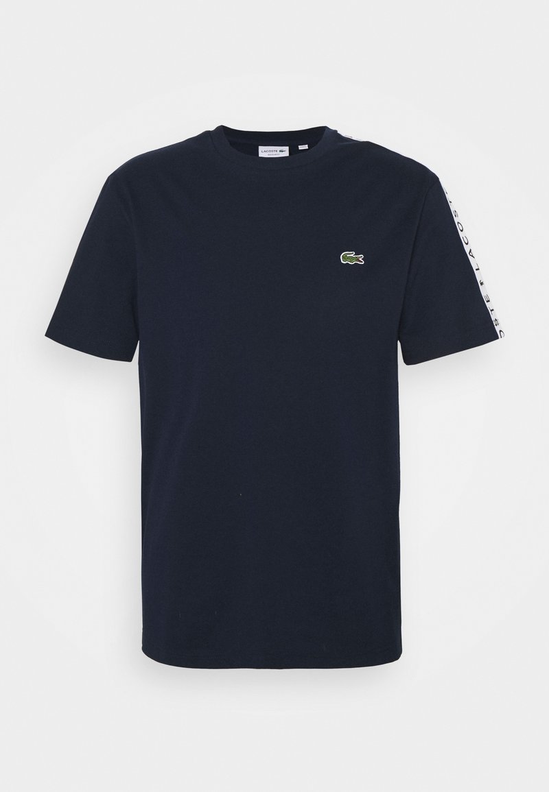 Marinblå bomulls t-shirt med rund hals, korta ärmar och en grön Lacoste-logotyp på bröstet. Vit textaccent vid sidans söm.