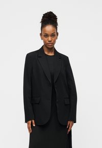 Μαύρο σακάκι με εφαρμοστή γραμμή, πέτα με εγκοπή, δύο εμπρόσθιες τσέπες και κούμπωμα με ένα κουμπί· φορεμένο πάνω από μαύρο τοπ. Ομαλή υφή υφάσματος.