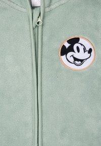 Giacca in pile verde chiaro con zip, caratterizzata da una toppa ricamata circolare di Topolino sul petto, con una texture liscia e morbida.