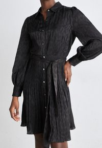 Vestido negro de botones con una cintura ajustada y falda plizada; presenta mangas abullonadas y un cinturón de lazo; patrón texturizado sutil en toda su extensión.