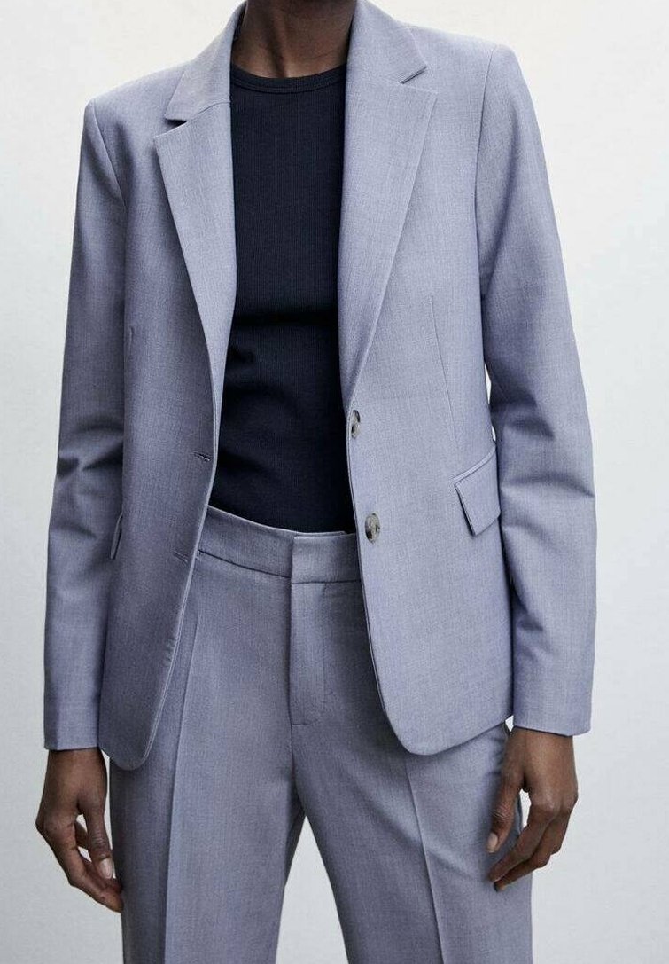 Personne portant un costume gris clair sur mesure avec un blazer à deux boutons et un pantalon assorti sur une chemise noire.