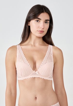 Donna che indossa un reggiseno in pizzo rosa chiaro con bordi smerlati e un piccolo fiocco al centro, mostrata dalla vita in su contro uno sfondo semplice.