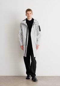 Parka impermeabil de culoare gri deschis, cu glugă, fermoar frontal și buzunare laterale, asortat cu pantaloni negri și pantofi. Textură netedă, design minimal.