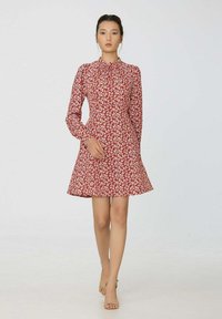 Femme marchant vers l'avant, portant une robe longue à manches longues florale rouge avec un col montant et des talons ouverts beiges sur un fond uni.