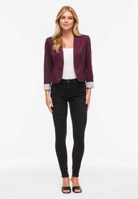 Blazer bordeaux à coupe ajustée, chemise blanche, jean skinny noir et sandales noires à enfiler ; détail de rayures sur les poignets des manches du blazer.