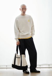 Cream sweatshirt with yellow logo, layered over yellow shirt, paired with dark pants. White and black canvas tote bag with label. Black sneakers.