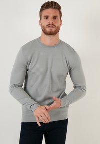 Maglione grigio a maglia con un motivo testurizzato a nido d'ape. Maniche lunghe e polsini e orlo a coste. Indossato con jeans scuri.