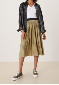 Camicia nera in pelle con bottoni, maglietta bianca a maniche lunghe, gonna midi khaki plissettata e sneakers verde oliva con suola bianca.