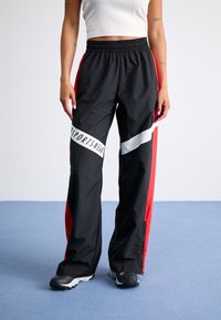 Black and red sports pants featuring an elastic waistband, side zippers, and a white logo stripe across the thigh. Smooth, lightweight material.