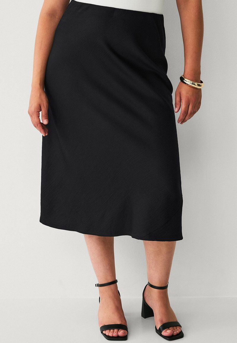 Zwarte midi-rok met een gladde textuur, gekenmerkt door een eenvoudig ontwerp en een lichte glans. Gecombineerd met zwarte hakken met enkelbandje.