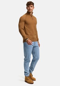 Bruine, gestructureerde pullover met een halve rits en geribde mouwen; gecombineerd met lichtblauwe denim jeans en beige wandelschoenen.