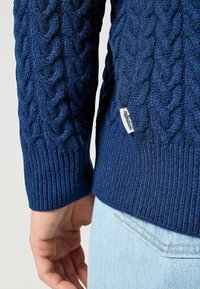 Granatowy sweter z dzianiny z wzorem w warkocze, ze ściągaczami przy mankietach i dolnym brzegu. Posiada małą białą metkę z napisem "Wrangler" na bocznym szwie.