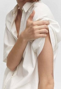 Chemise blanche ample à boutons avec manches retroussées. Tissu doux et léger avec une finition légèrement texturée. Le bras découvert montre un petit tatouage.