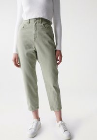 Světle zelené džíny s vysokým pasem a volným střihem, se pěti kapsami a zapínáním na knoflík, v kombinaci s bílými teniskami na jednobarevném pozadí.