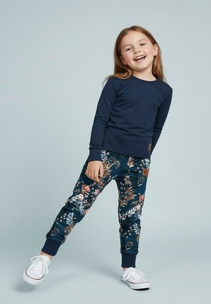 Uśmiechnięte dziecko pochylające się, ubrane w granatową bluzę z długim rękawem, spodnie w kwiatowy wzór oraz białe trampki na jednolitym tle.