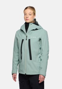 Giacca impermeabile verde chiaro con chiusura a zip, cappuccio regolabile e dettagli neri. Presenta due tasche frontali verticali e materiale interno testurizzato.