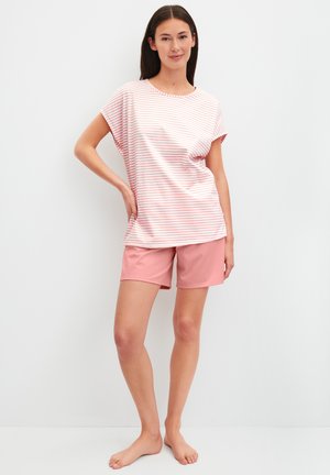 Frau steht barfuß, trägt ein rosa-weiß gestreiftes kurzärmliges Oberteil und einfarbige rosa Shorts vor einem schlichten weißen Hintergrund.